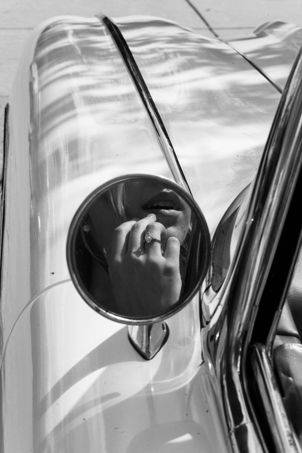 Black and white photo of a person's reflection in a vintage car's round side mirror, wearing a ring.