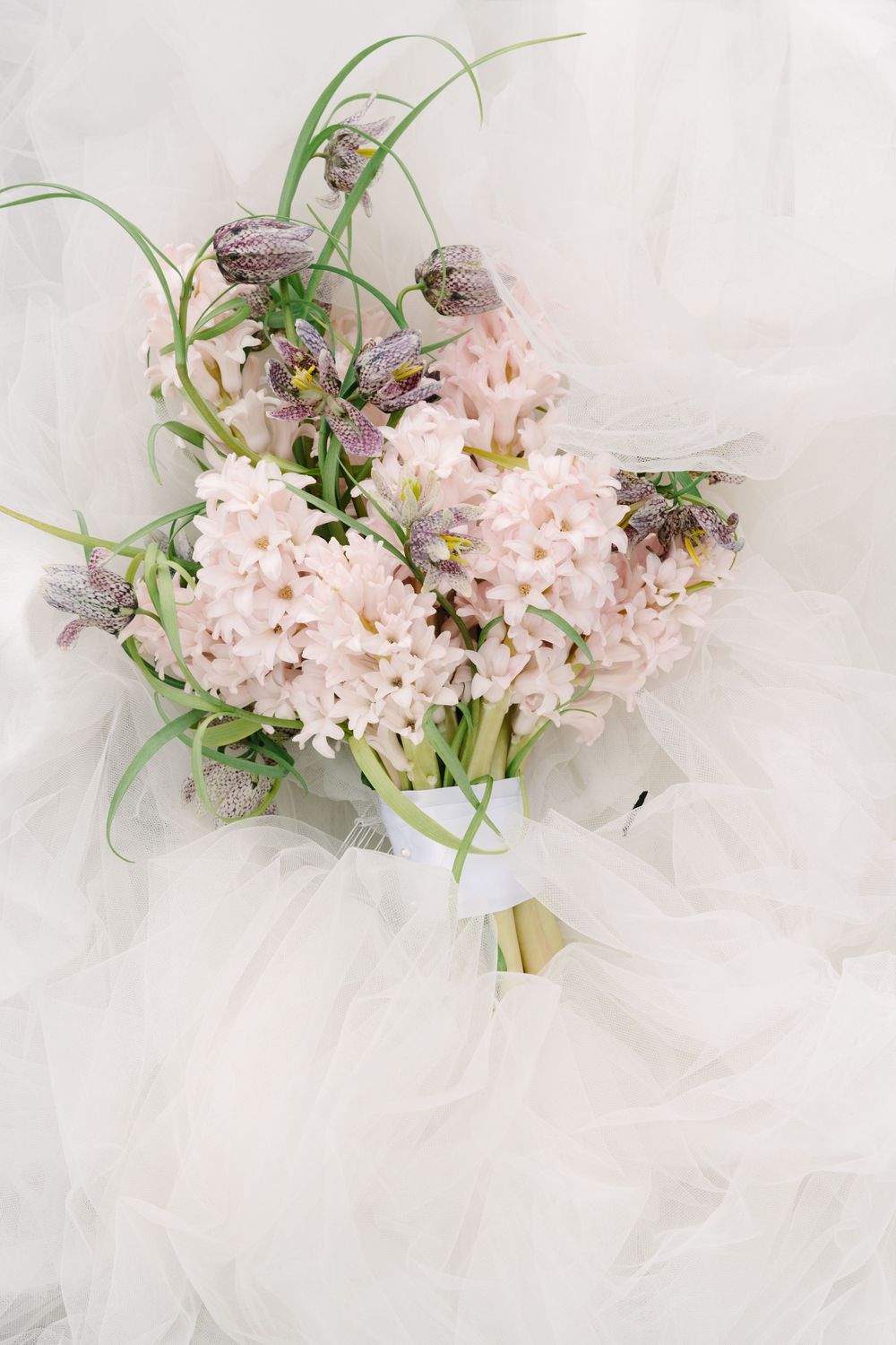 Elegant bridal bouquet of pale pink hyacinths and purple fritillaria flowers tied with white ribbon on tulle fabric.