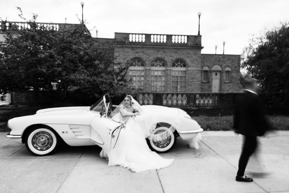 Black and white wedding photo of bride in vintage Corvette convertible with groom approaching, elegant building backdrop.