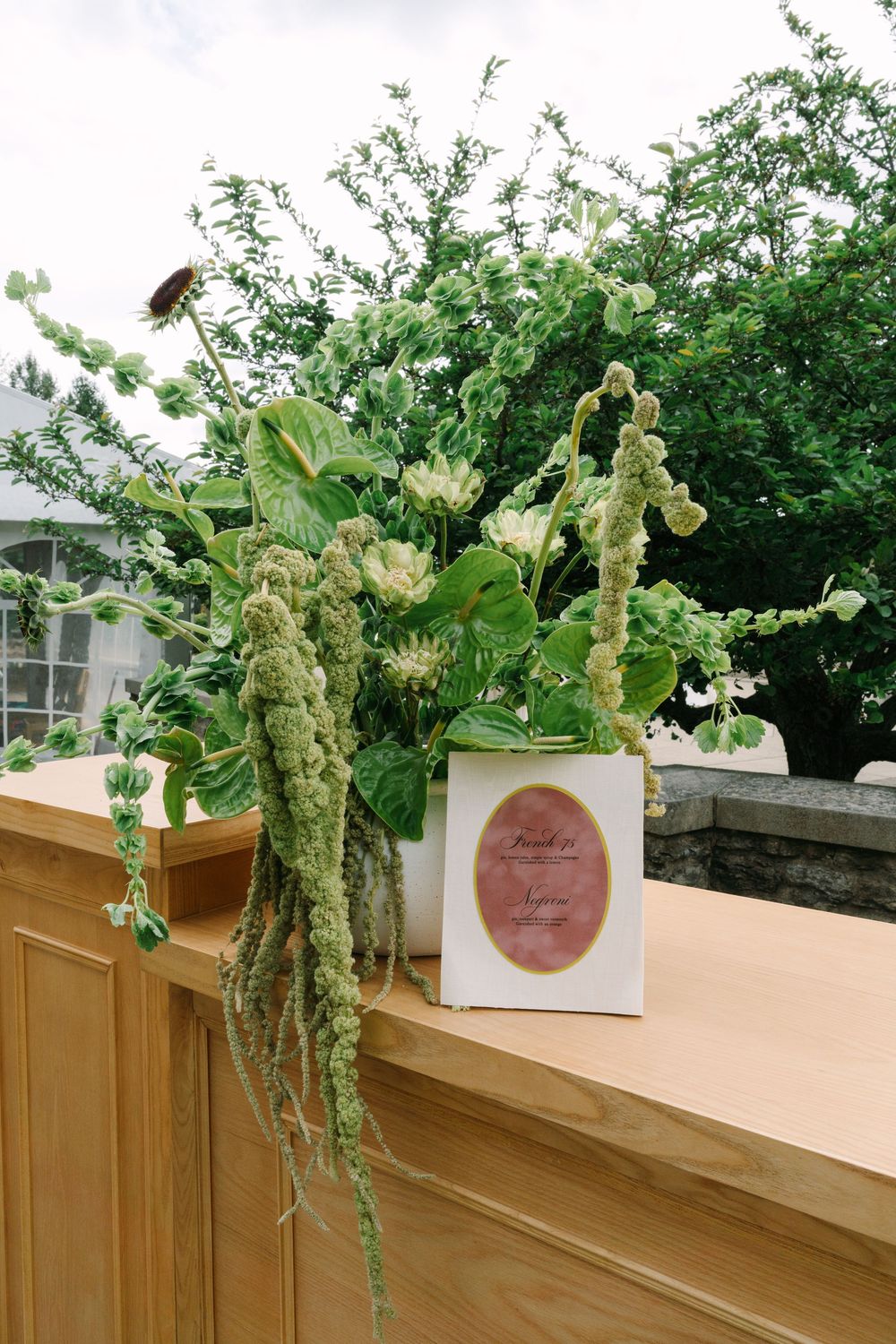 White box with pink oval label sits on wooden ledge adorned with lush green botanical arrangement at outdoor wedding.