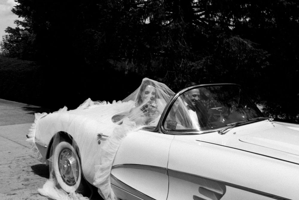 Bride in wedding dress posing beside a classic vintage convertible sports car in black and white photo.
