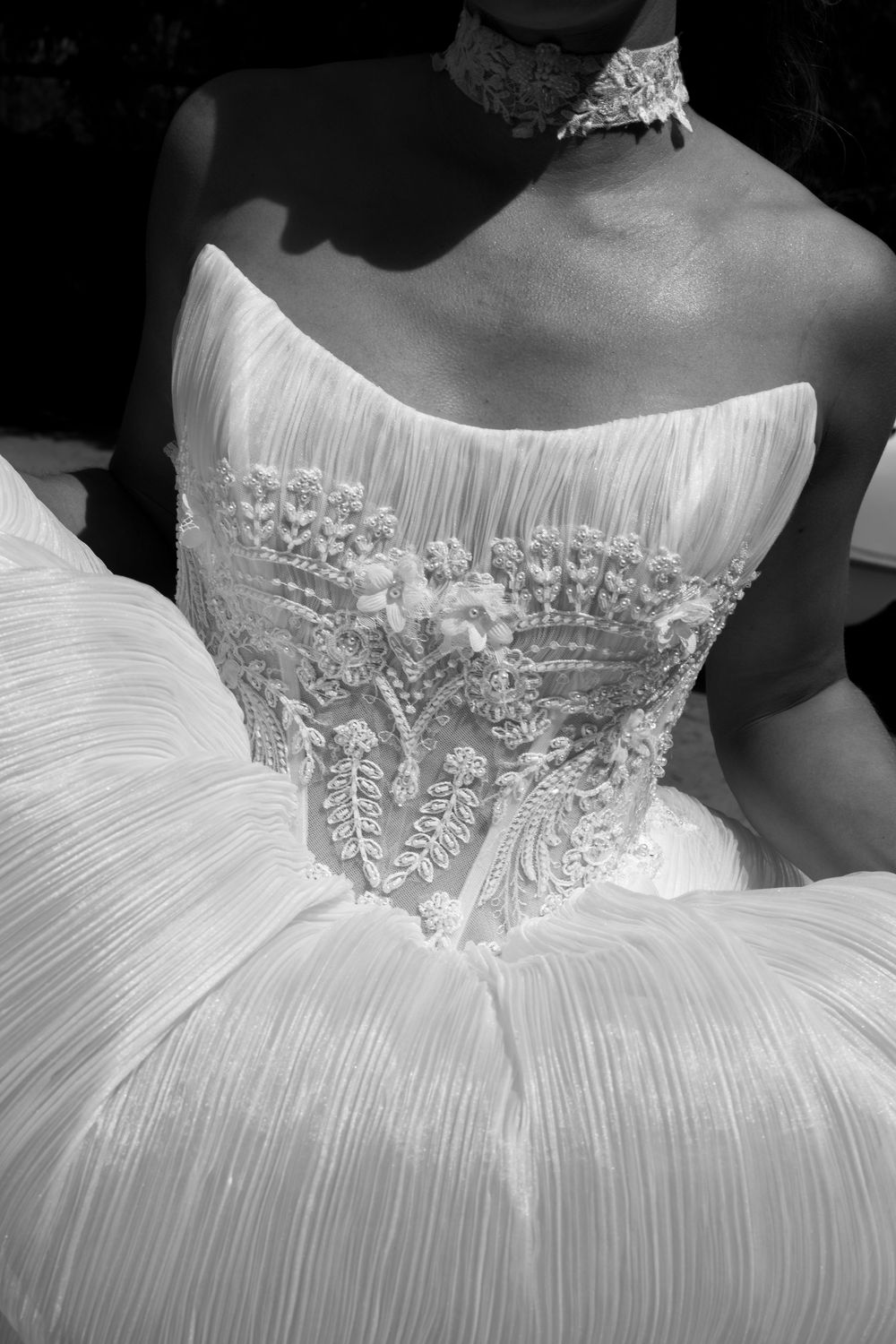 Black and white close-up of a strapless bridal gown with intricate lace bodice embroidery and pleated skirt detailing.