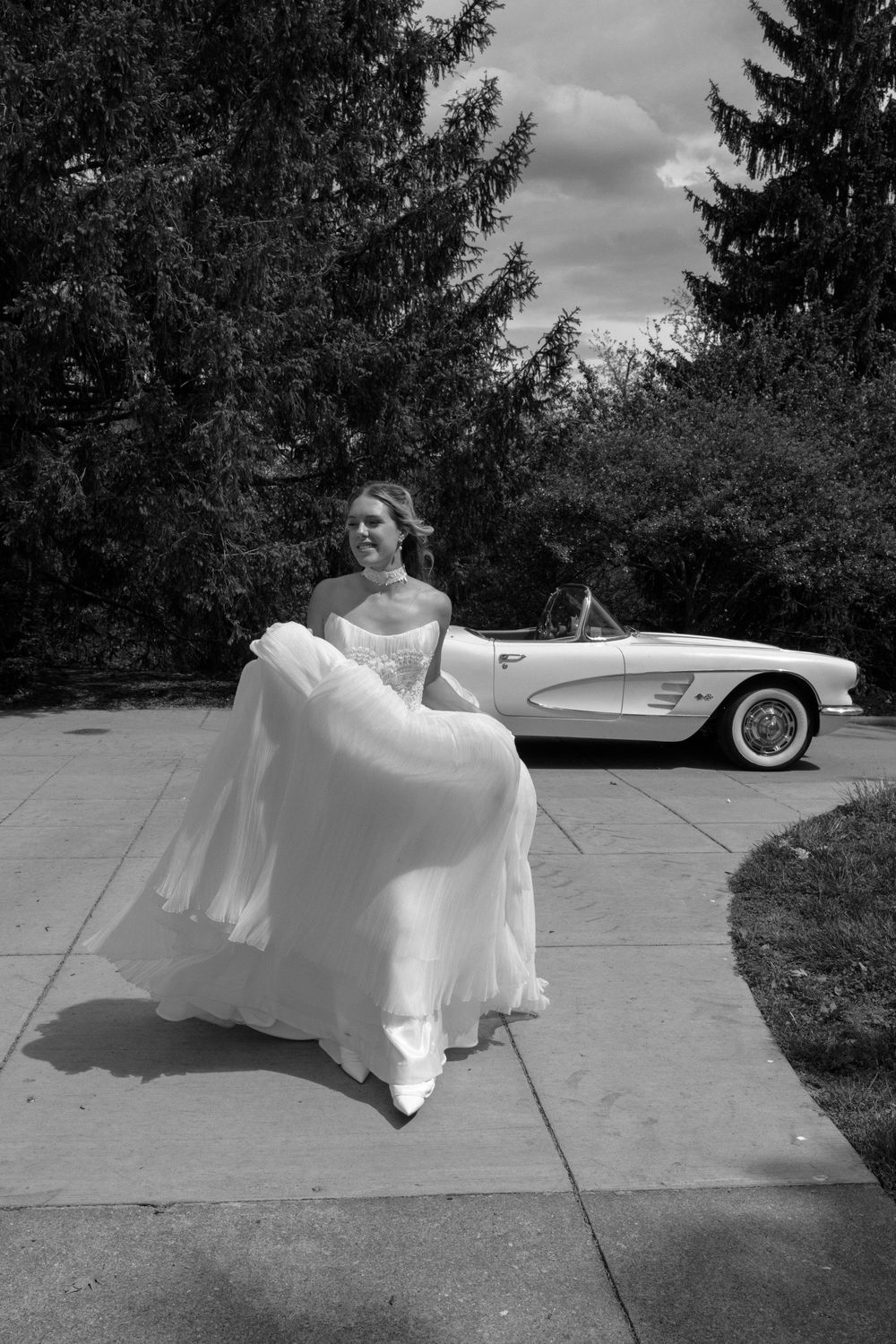 Joyful bride in flowing white gown lifts her dress while standing near a classic white Corvette convertible outdoors.