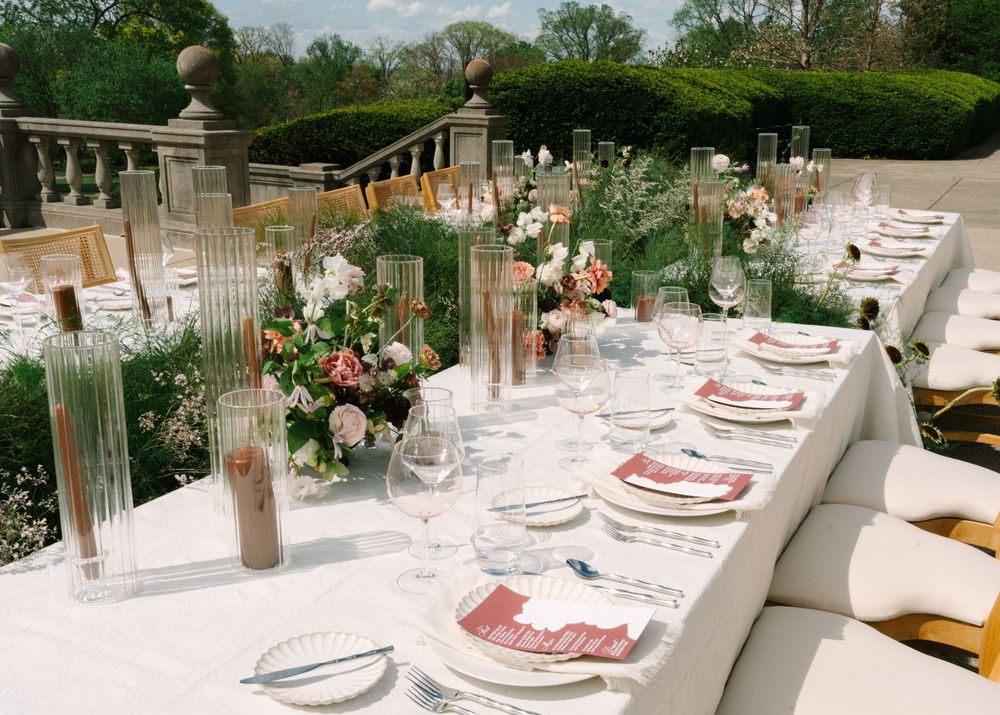 Elegant outdoor wedding reception table with floral centerpieces, crystal glassware, and pink napkins at a garden venue.