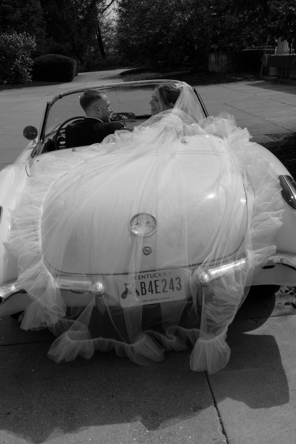 Bride and groom in a vintage white Corvette convertible with wedding veil draped over the car, Kentucky license plate B4E243.