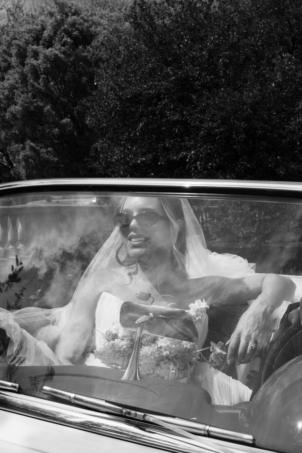 Black and white photo of a bride in sunglasses and veil sitting in a vintage convertible car holding a bouquet.