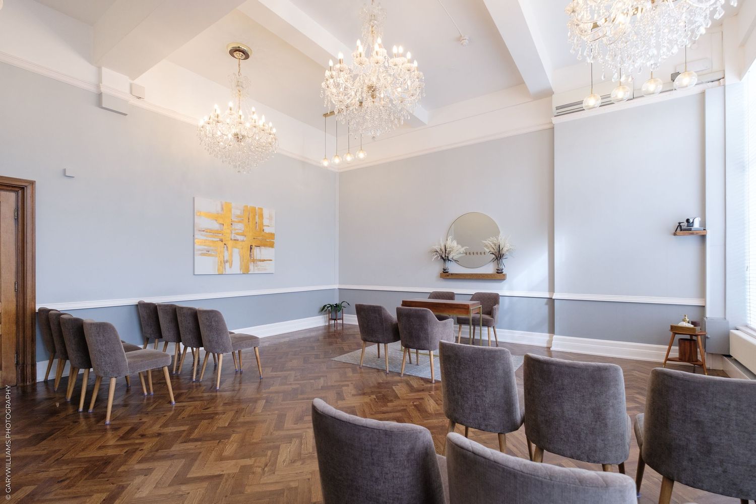 Elegant event room with crystal chandeliers, gray velvet chairs, parquet flooring, and gold abstract artwork on gray walls.