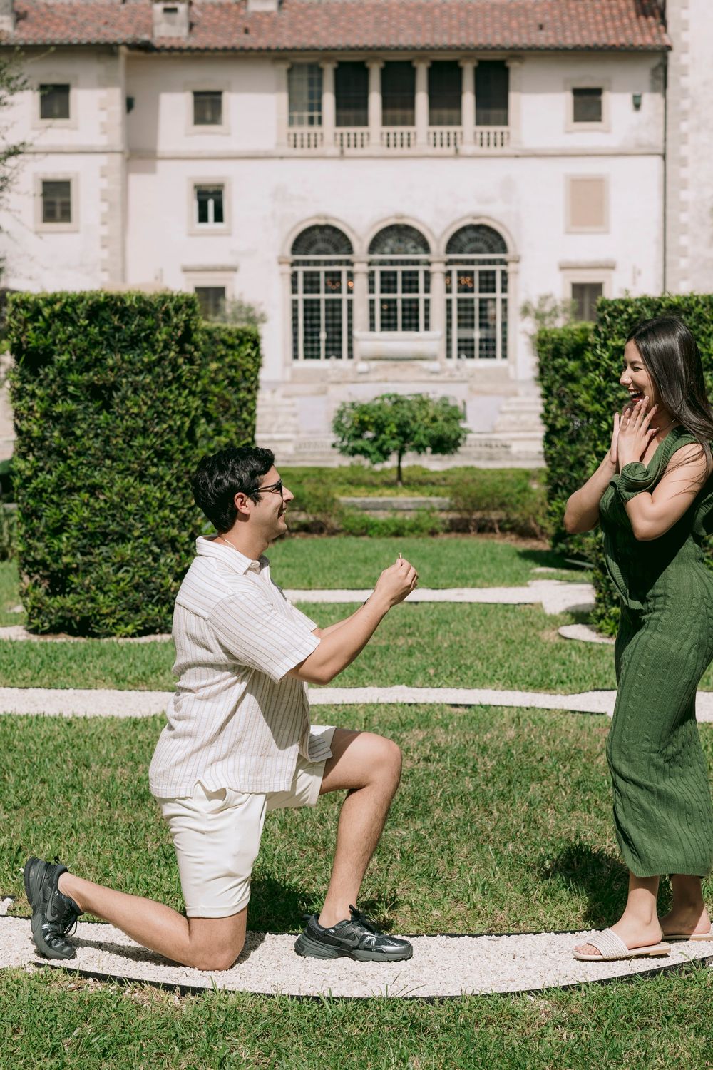 Man proposes on one knee to woman in green dress in lush garden outside elegant white villa.