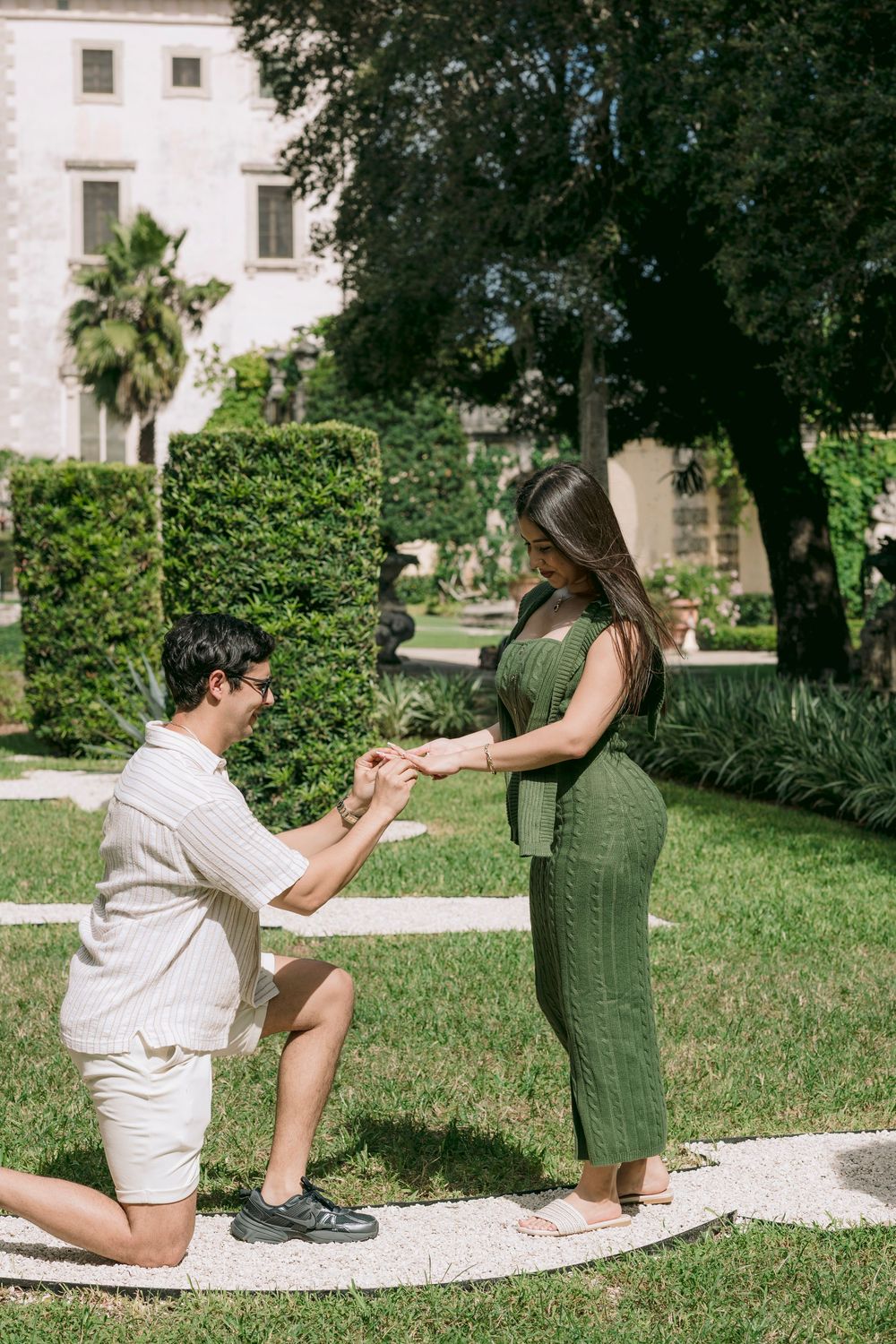 Man proposing to woman in green dress in a lush garden near a historic building on a sunny day.