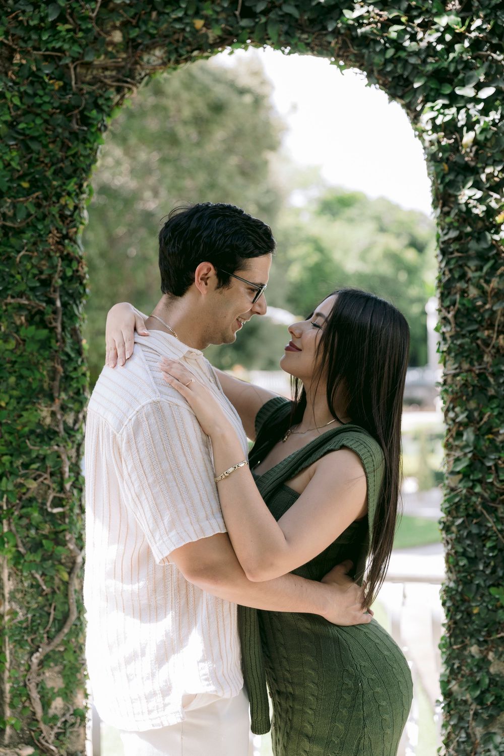 Romantic couple embracing and smiling under an ivy-covered stone arch in a garden.