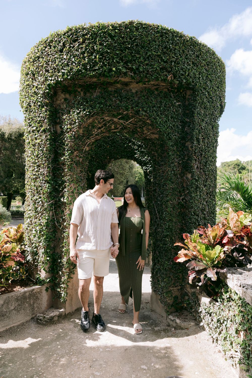 Couple holding hands walking through ivy-covered stone archway in lush tropical garden.