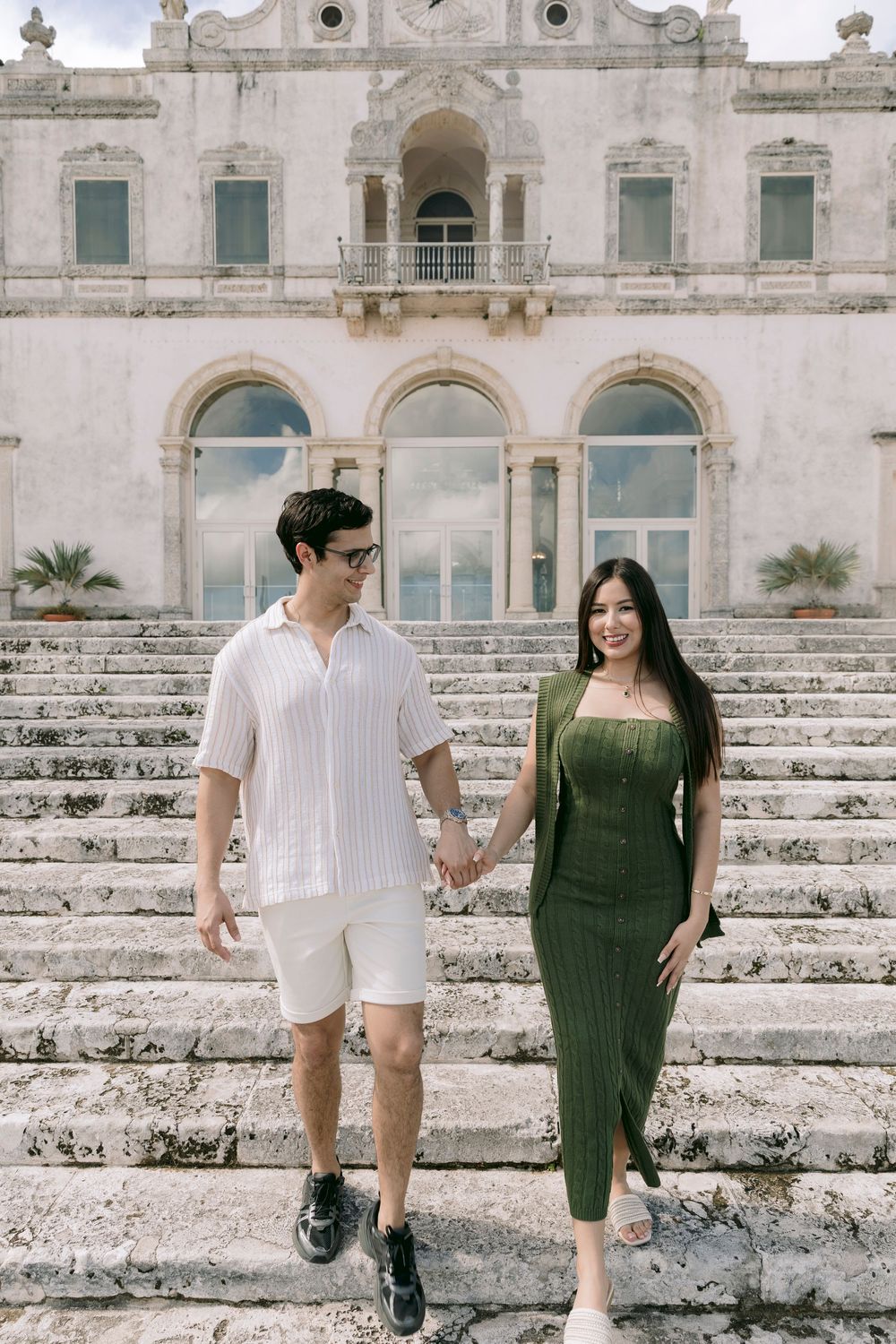 Couple holding hands on ornate stone steps of historic villa facade.