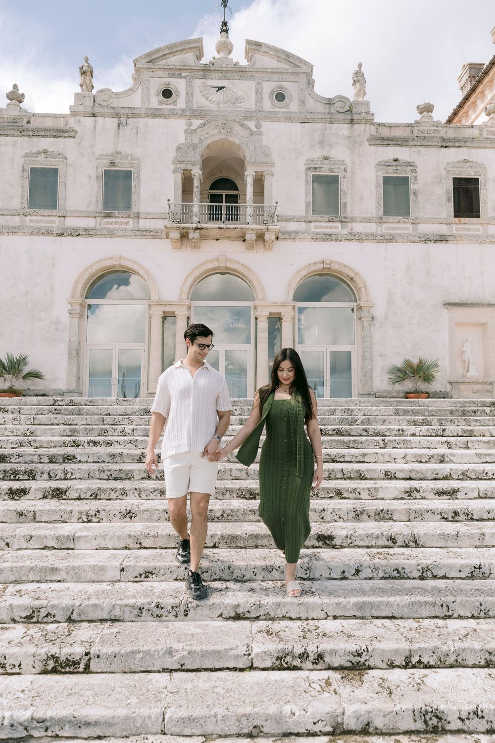 Couple holding hands walking down grand stone steps of historic European villa.