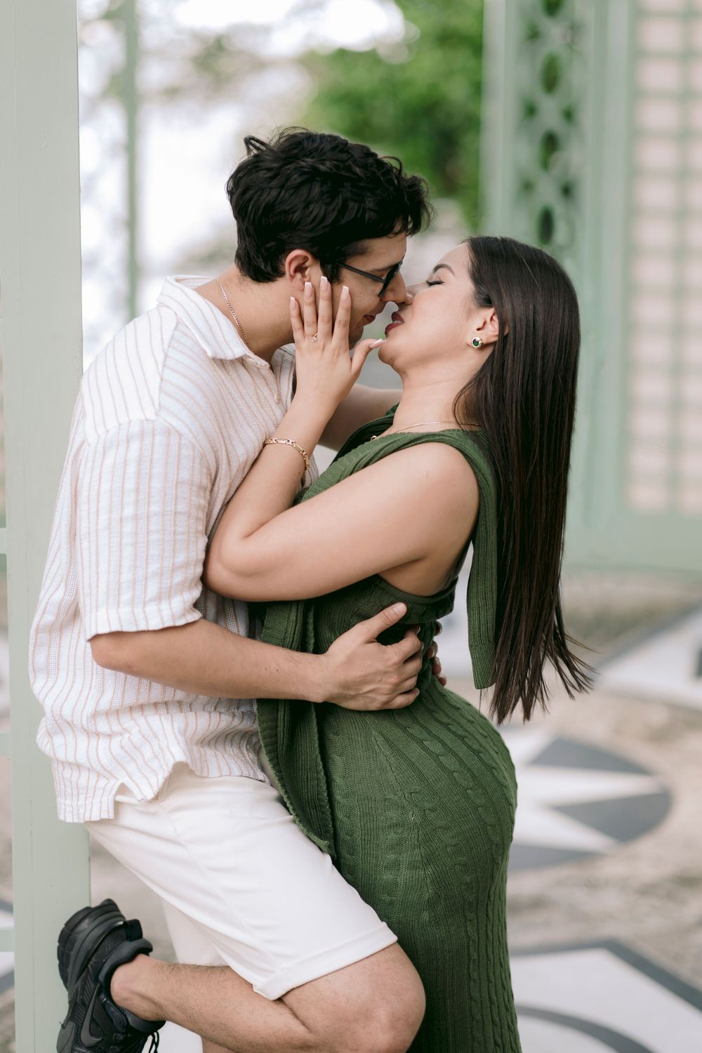 A couple sharing a romantic kiss outdoors, with the woman in a green dress and man in a striped shirt embracing.