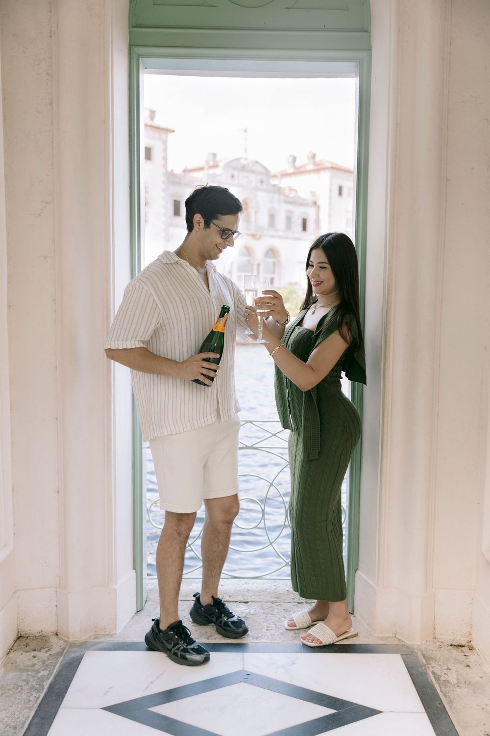 Couple toasting champagne in a doorway overlooking a Venice canal, man in shorts and woman in green dress.
