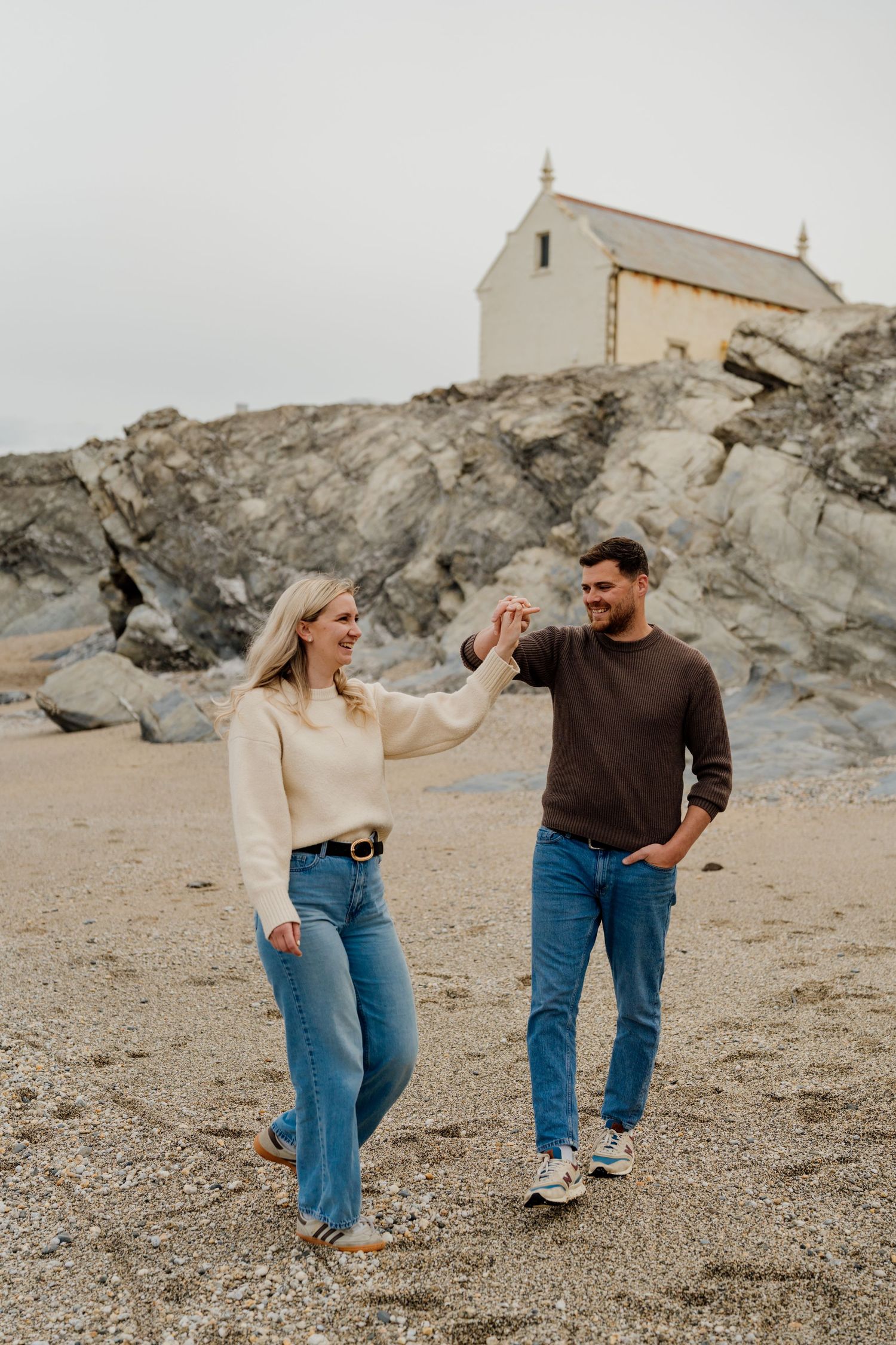 Couple holding hands on a rocky beach with a white boathouse in the background.