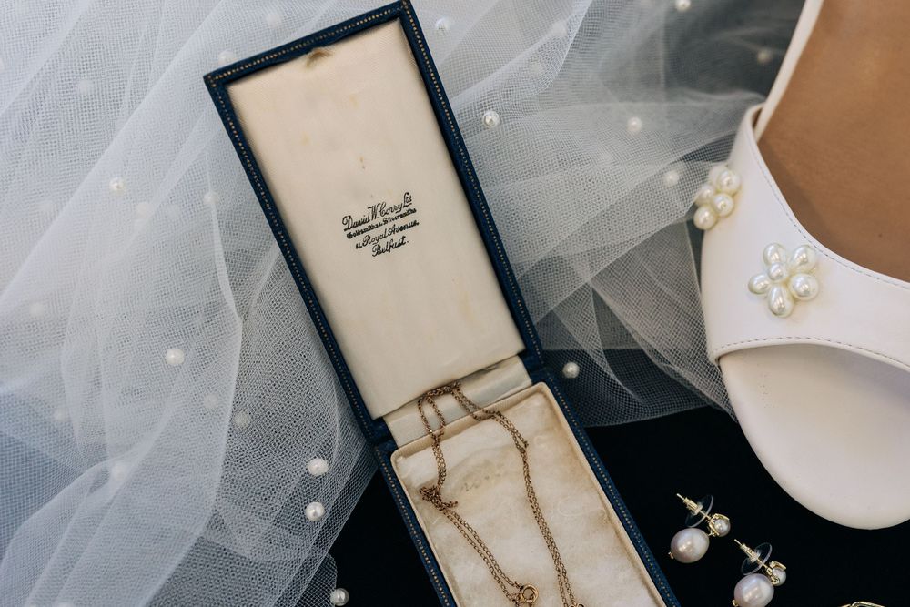 Vintage gold necklace in navy jewelry box beside pearl earrings and white bridal heel with veil.