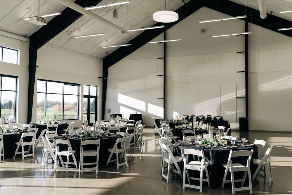 Elegant wedding reception hall with black and white decor, round tables, white chairs, and large windows in a modern venue.