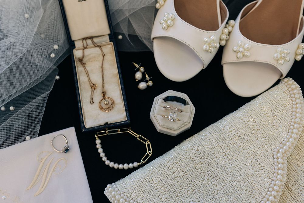 Elegant bridal accessories flatlay featuring pearl shoes, jewelry, clutch bag, and veil on dark background.