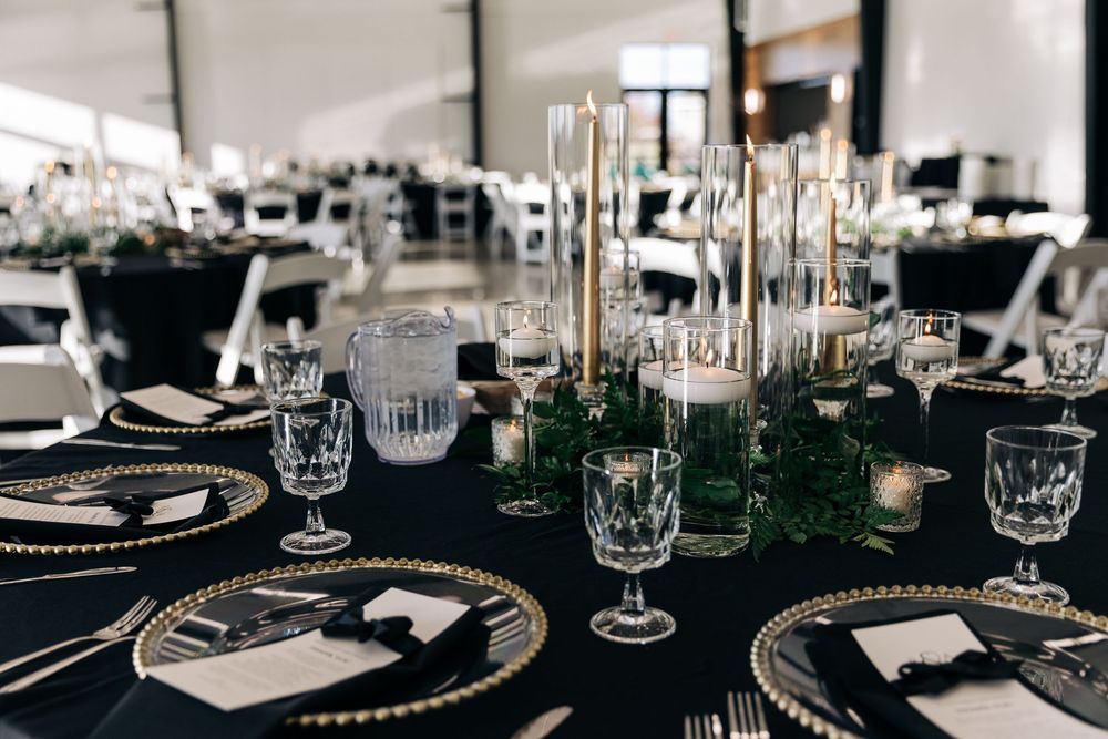 Elegant black and gold wedding reception table with crystal glassware, tall candles, and lush greenery centerpiece.