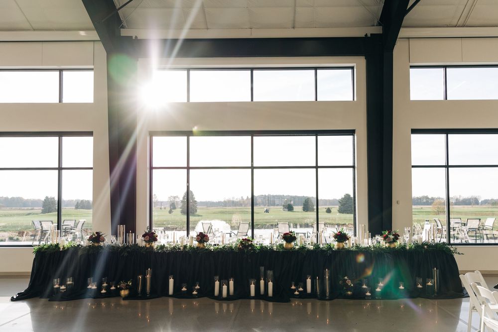 Elegant wedding head table with dark linen, floral centerpieces, and candles before large windows overlooking green fields.