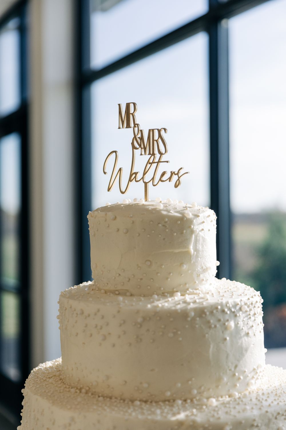 Three-tier white wedding cake with pearl details and gold Mr. & Mrs. Walters cake topper near large windows.
