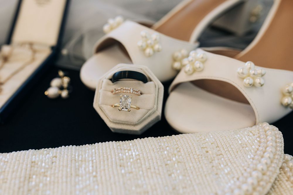 Elegant bridal flat lay featuring diamond rings in a velvet box, pearl-embellished white heels, and delicate jewelry.
