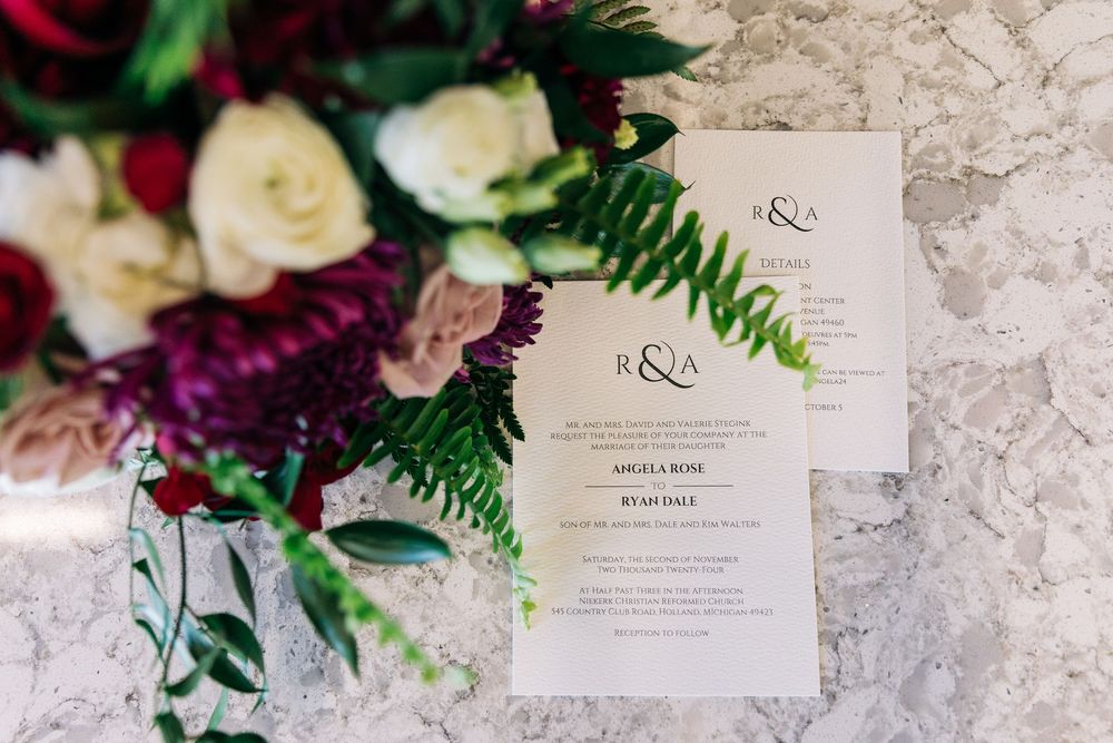 Elegant wedding invitation suite with fern greenery beside a lush bouquet of burgundy and cream flowers on marble.