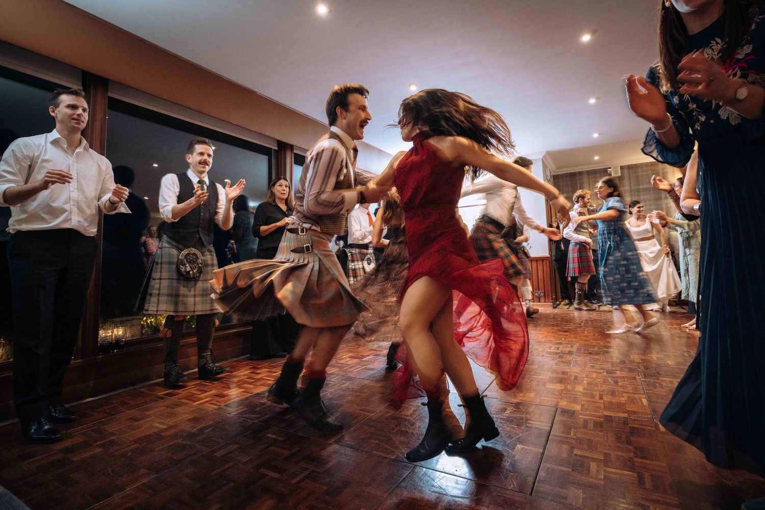 Energetic Scottish ceilidh dancers in traditional kilts and red dress spin on a wooden dance floor at a lively wedding reception.