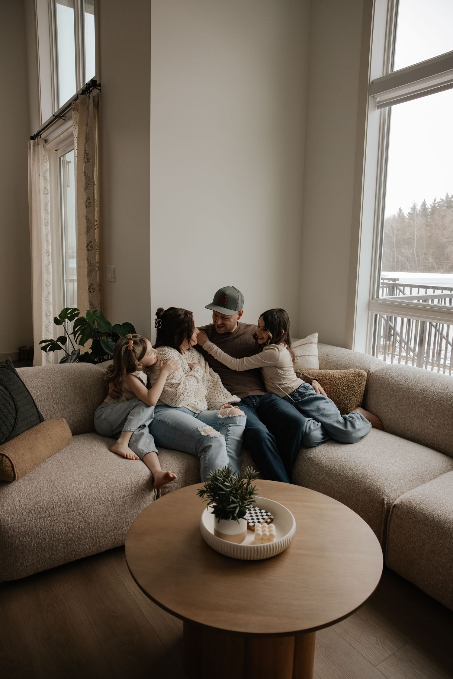 Family of four cuddling on a beige sectional sofa in a modern living room with large windows and a round wood coffee table.