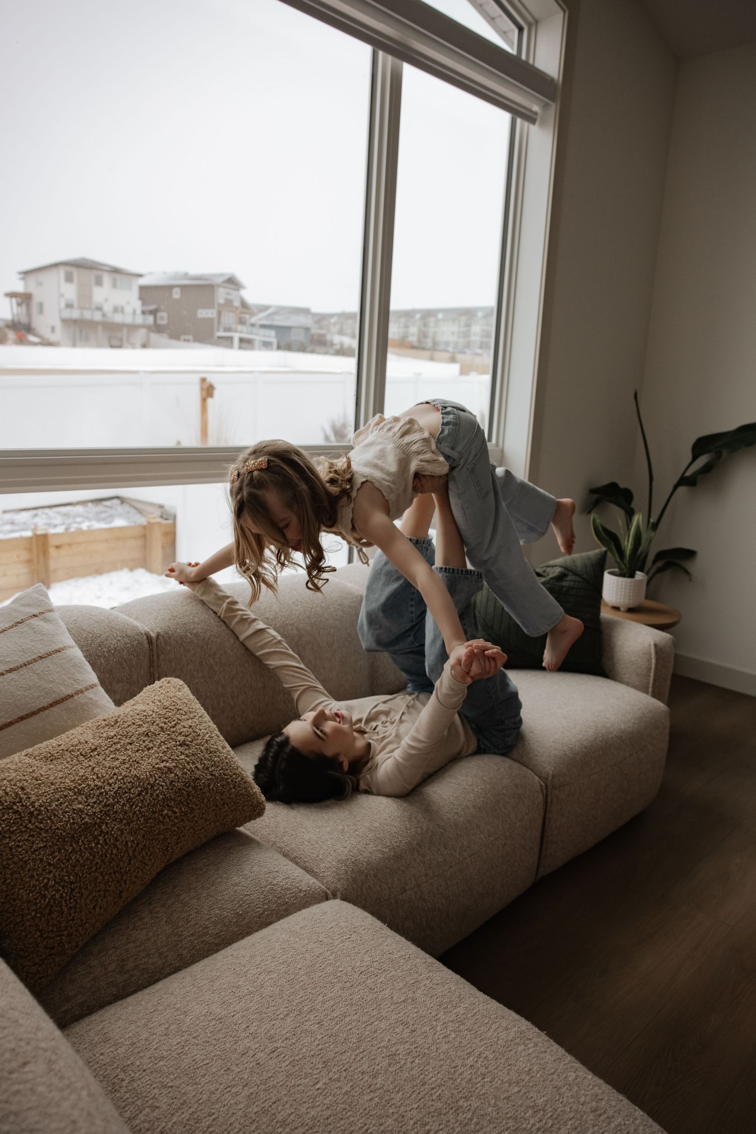 Parent lying on couch lifting child in playful indoor family moment with snowy window view.
