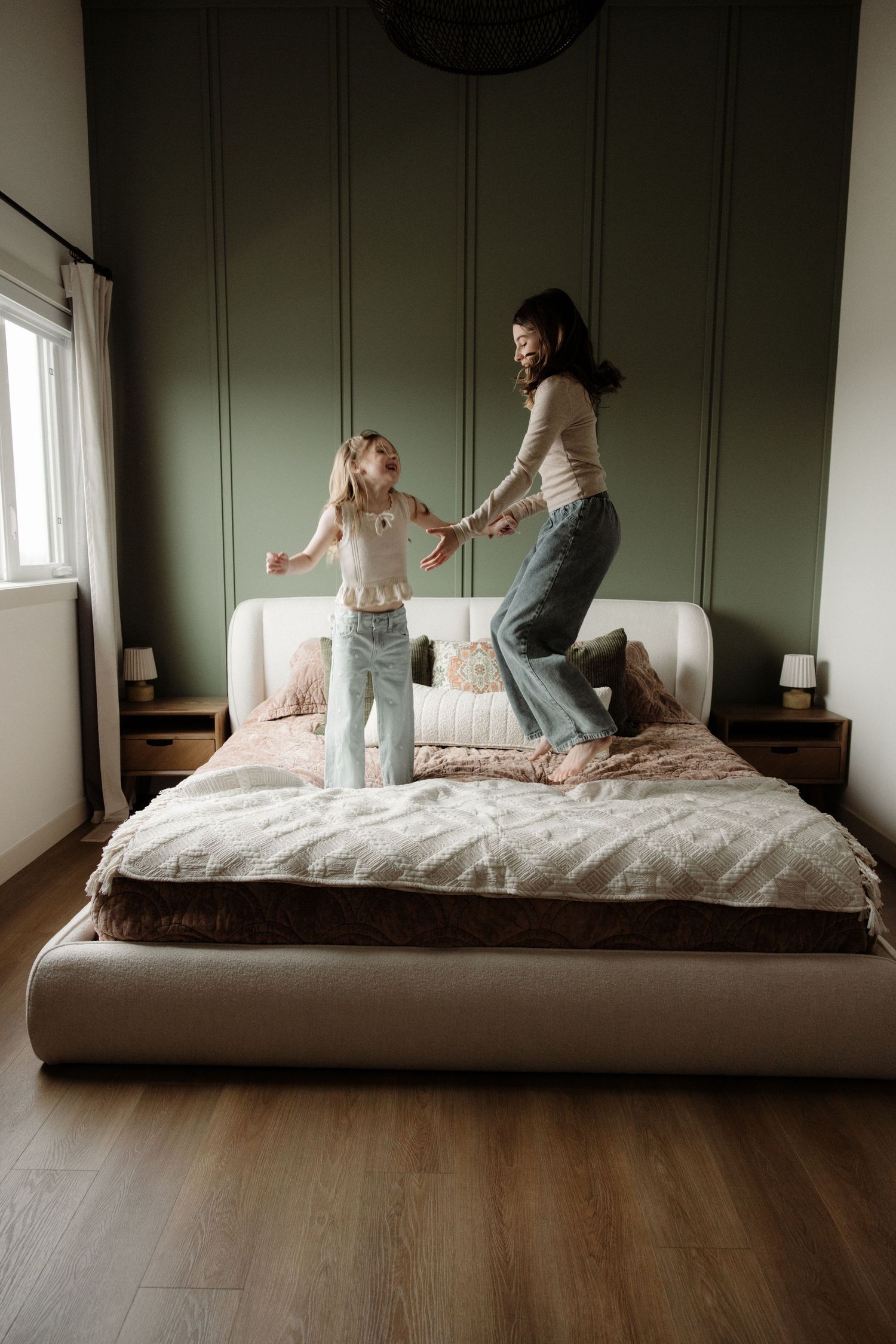 Mother and daughter joyfully jumping on a bed in a stylish green bedroom.