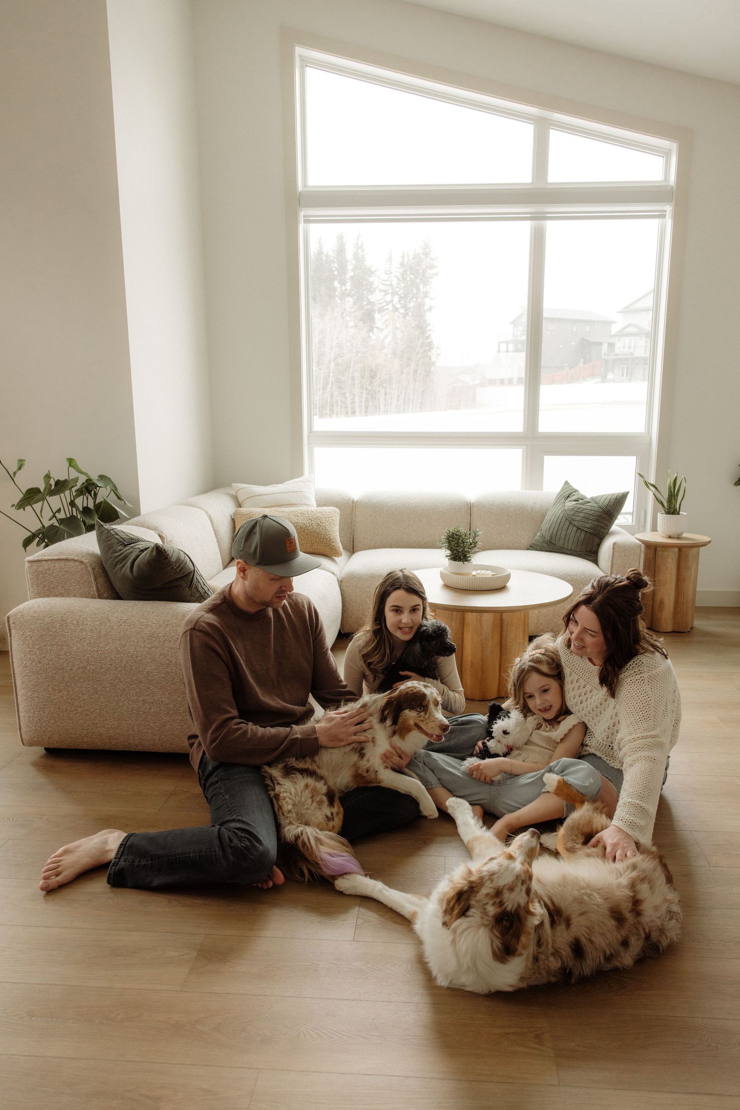 Family sitting on living room floor with large dog, enjoying time together near bright window and modern sofa.