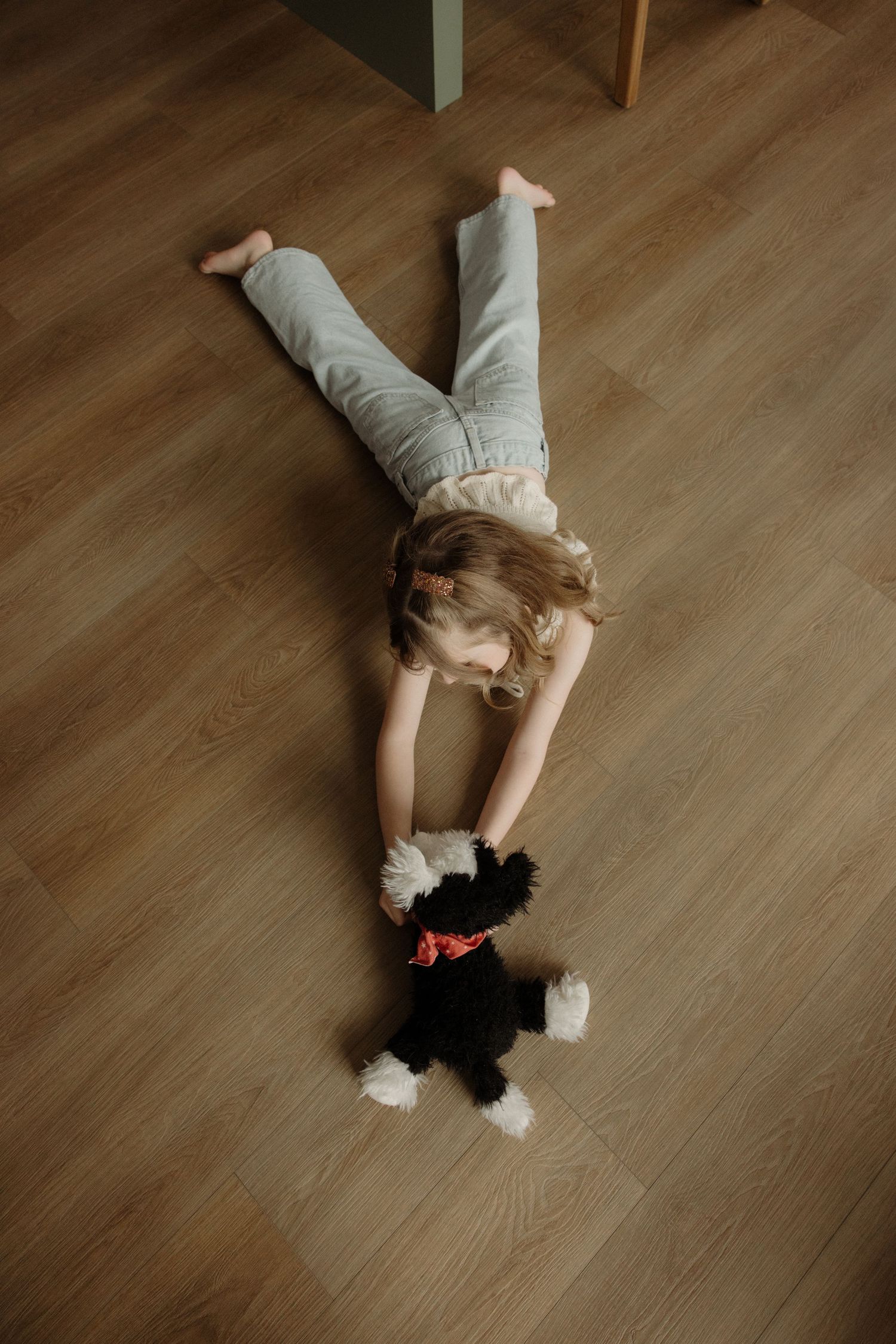 Young girl lying on hardwood floor playing with a black and white stuffed animal toy.