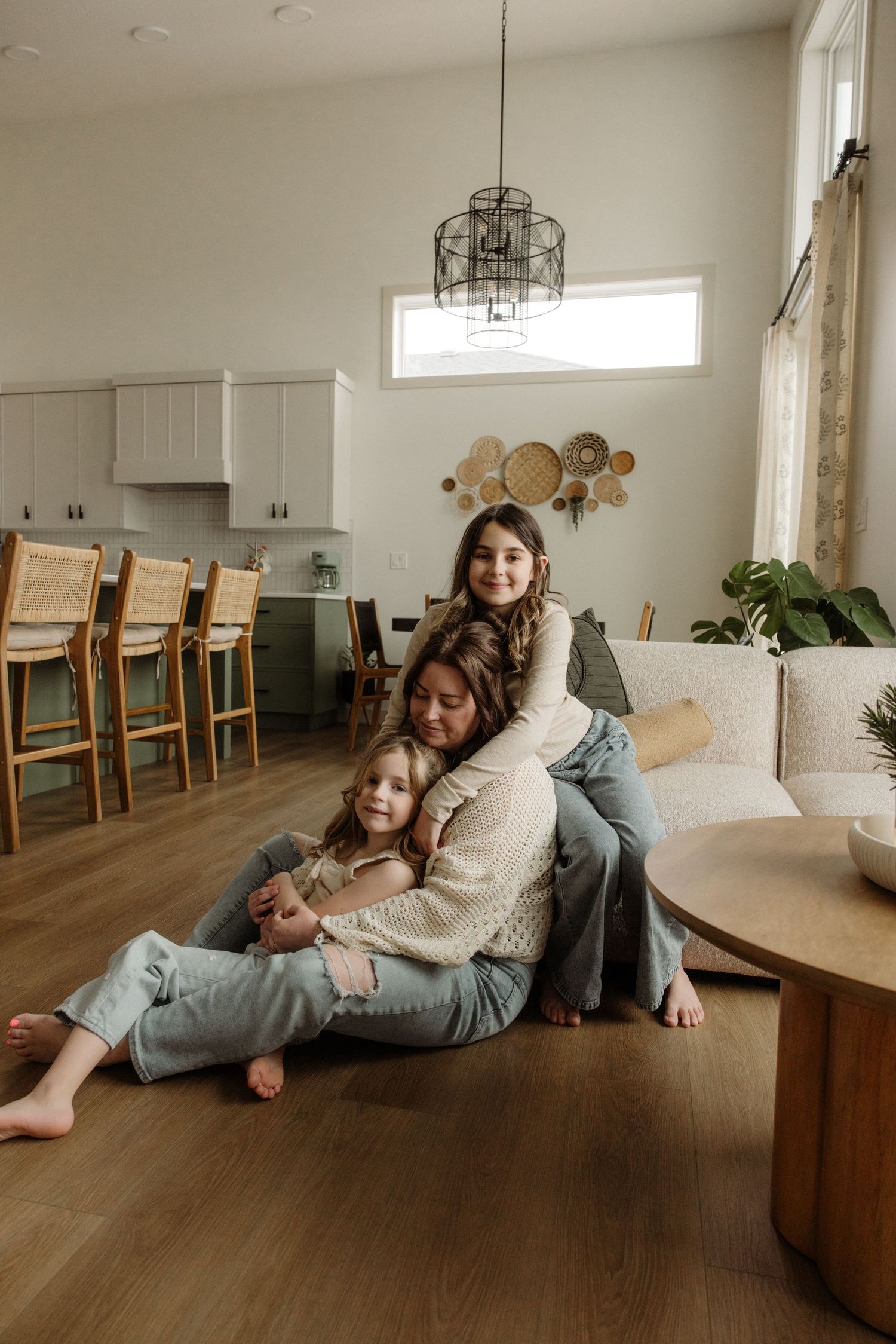 Mother and two daughters hugging on floor of bright open-concept living room with wood floors.