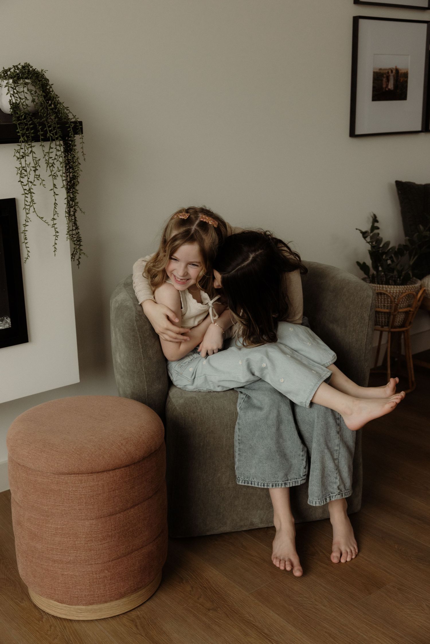Two sisters cuddling and laughing together in a gray armchair in a cozy living room.
