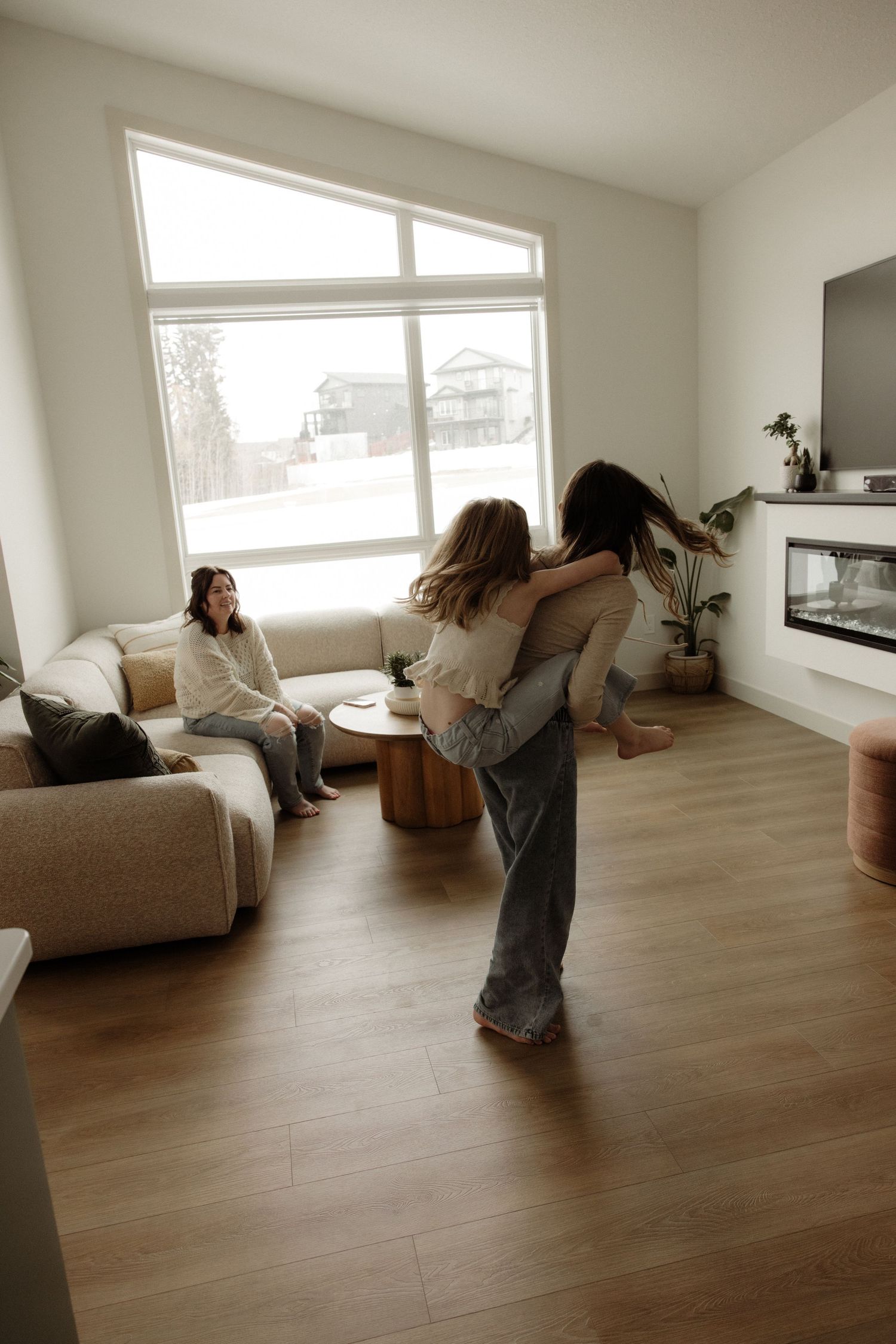 Adult lifting child in joyful hug in modern living room while another person watches from couch.