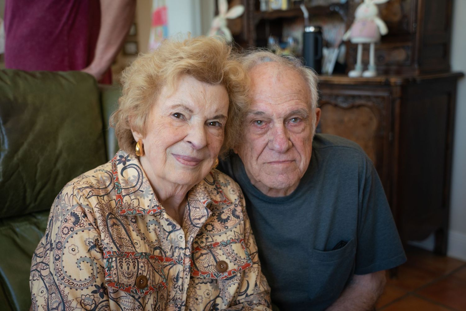 Elderly couple smiling warmly together indoors, woman in floral top and man in gray shirt, seated closely.