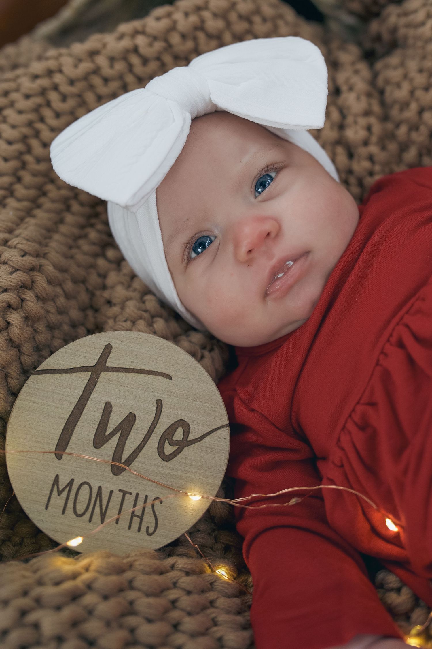 A two-month-old baby girl in a red outfit and white bow headband lies on a knit blanket with a wooden milestone sign.