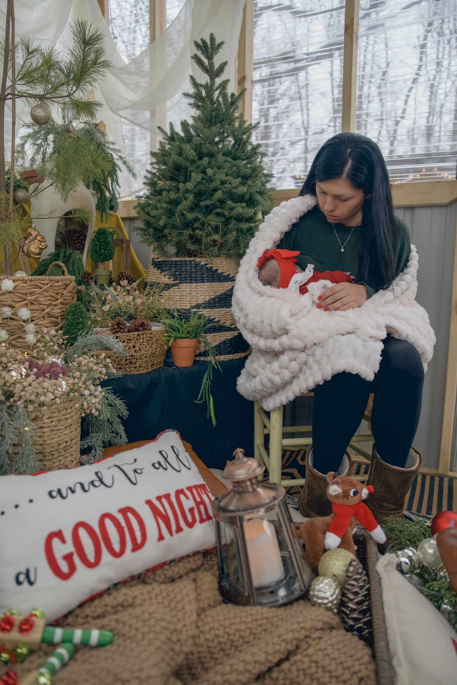 Mother cradling newborn in white blanket surrounded by Christmas decorations and trees in cozy holiday setting.