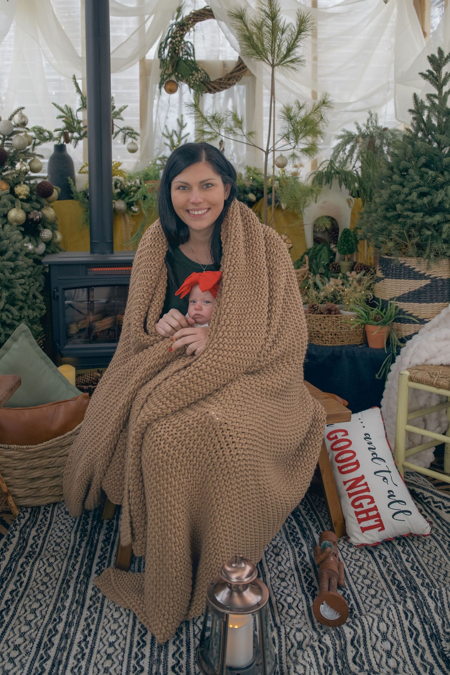 Woman wrapped in cozy knit blanket holding newborn baby by Christmas tree and wood stove holiday decor.