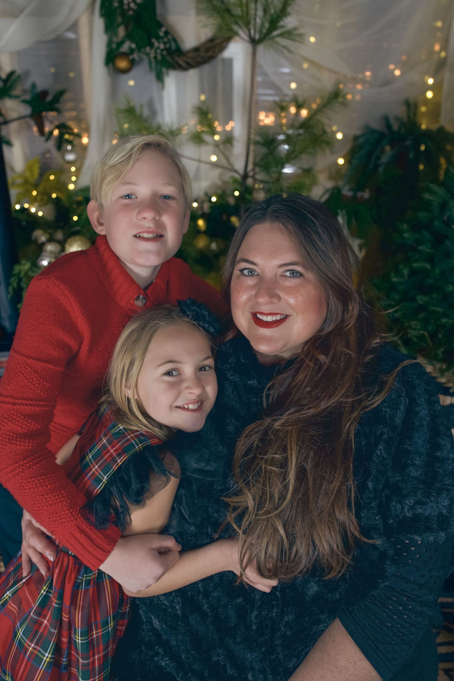 A woman and two children smile together in front of festive Christmas lights and greenery, wearing holiday outfits.