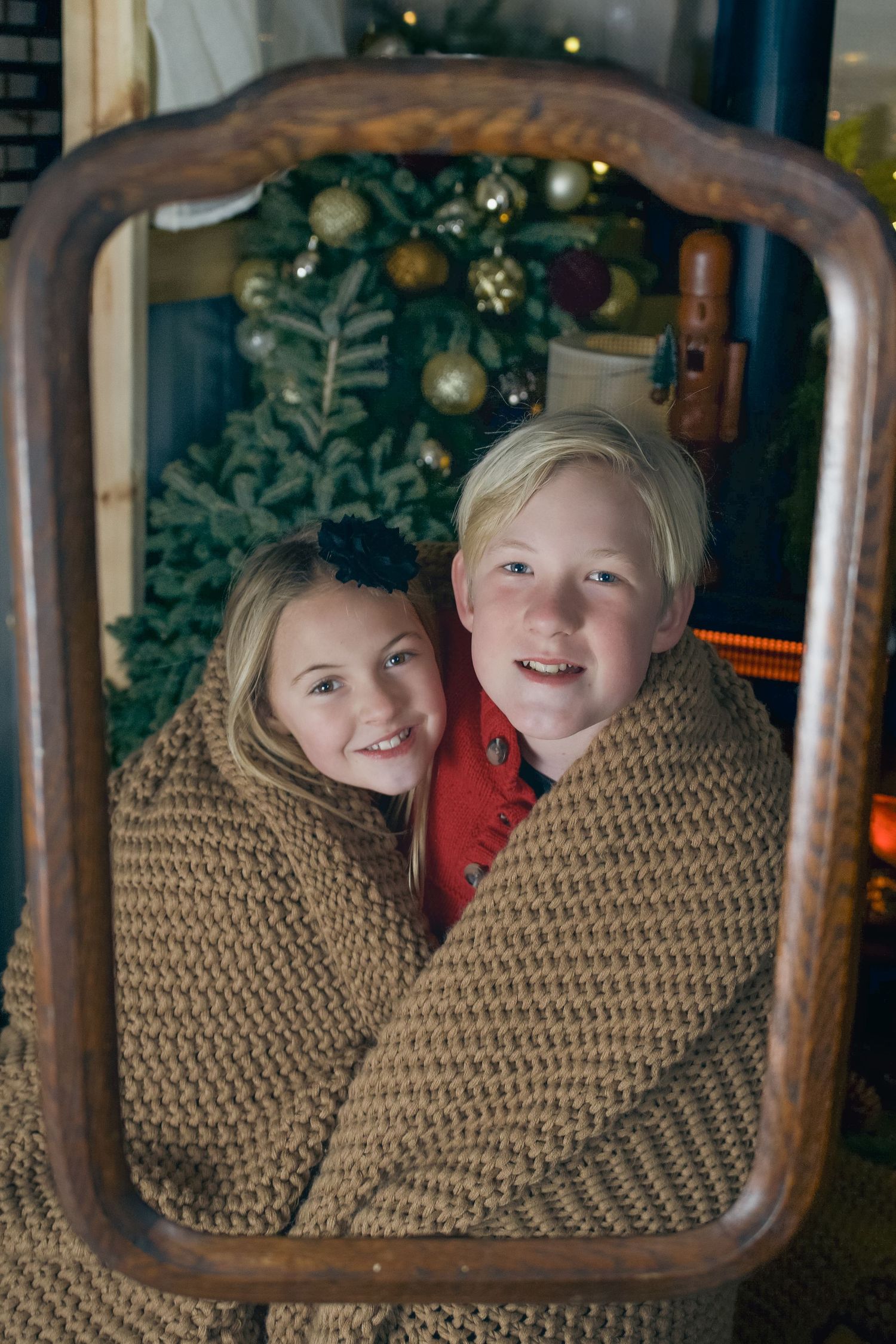 Two children wrapped in a knit blanket smile together in front of a decorated Christmas tree.