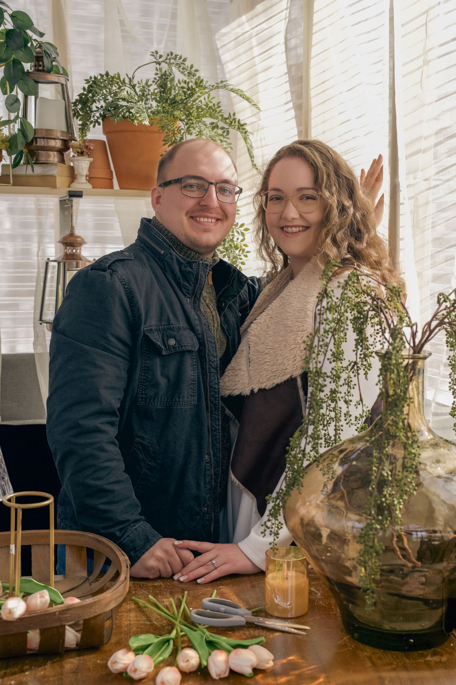 A smiling couple poses together among lush plants and tulips on a wooden table in a cozy botanical setting.