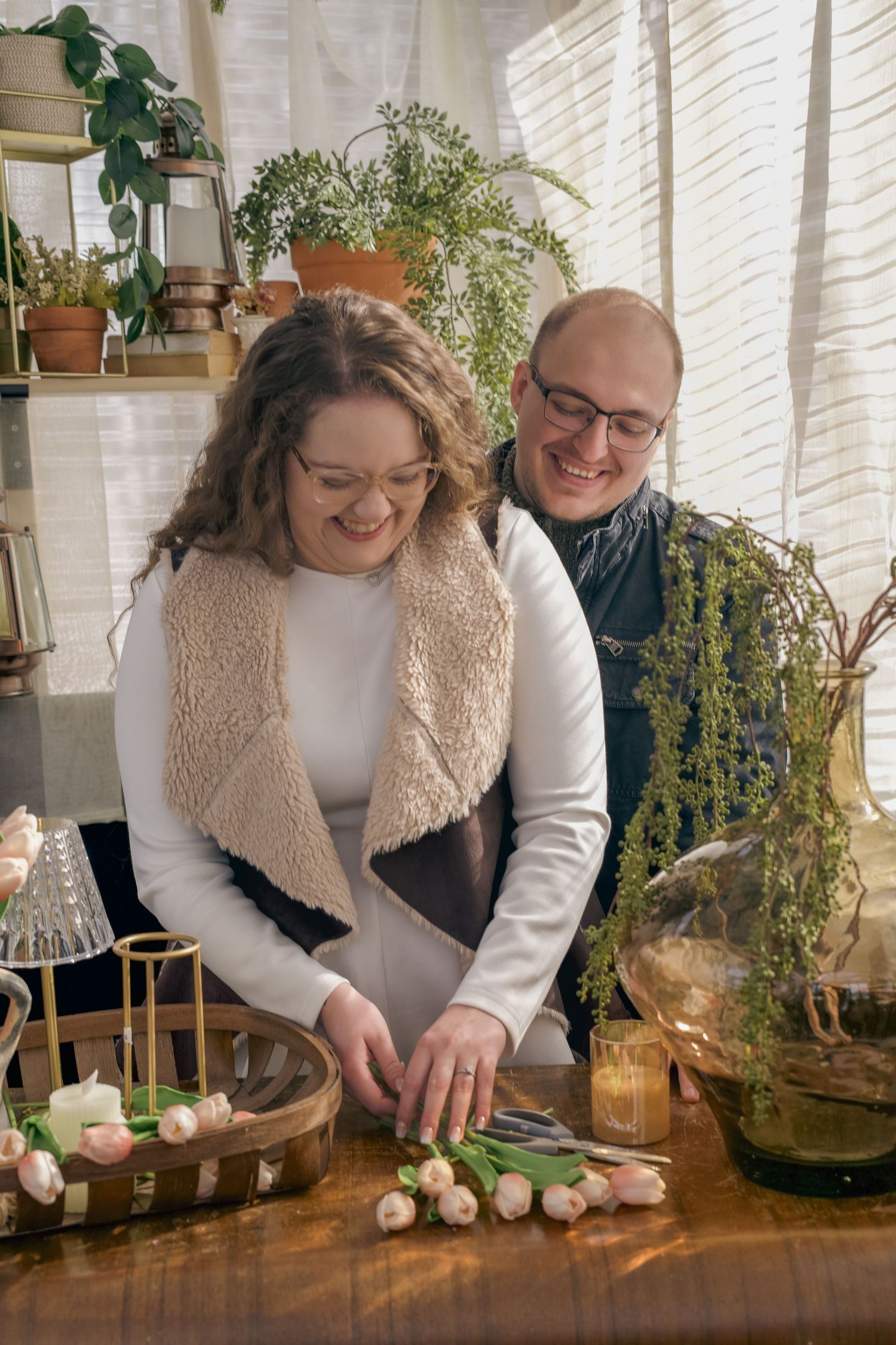 A smiling couple arranges tulips on a wooden table in a cozy plant-filled room.