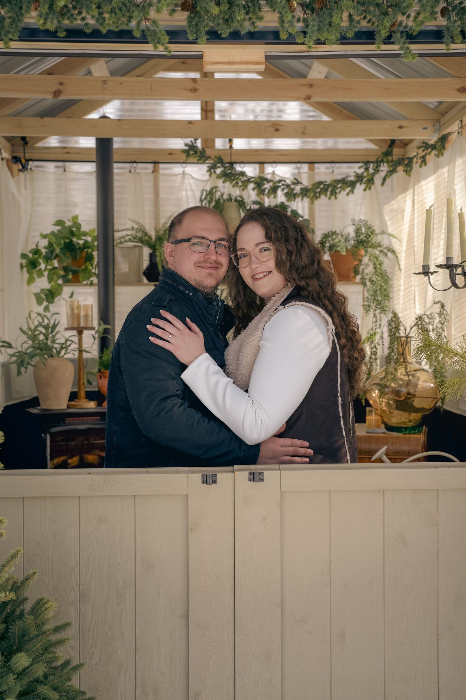 A couple embraces warmly inside a cozy greenhouse decorated with plants and greenery.