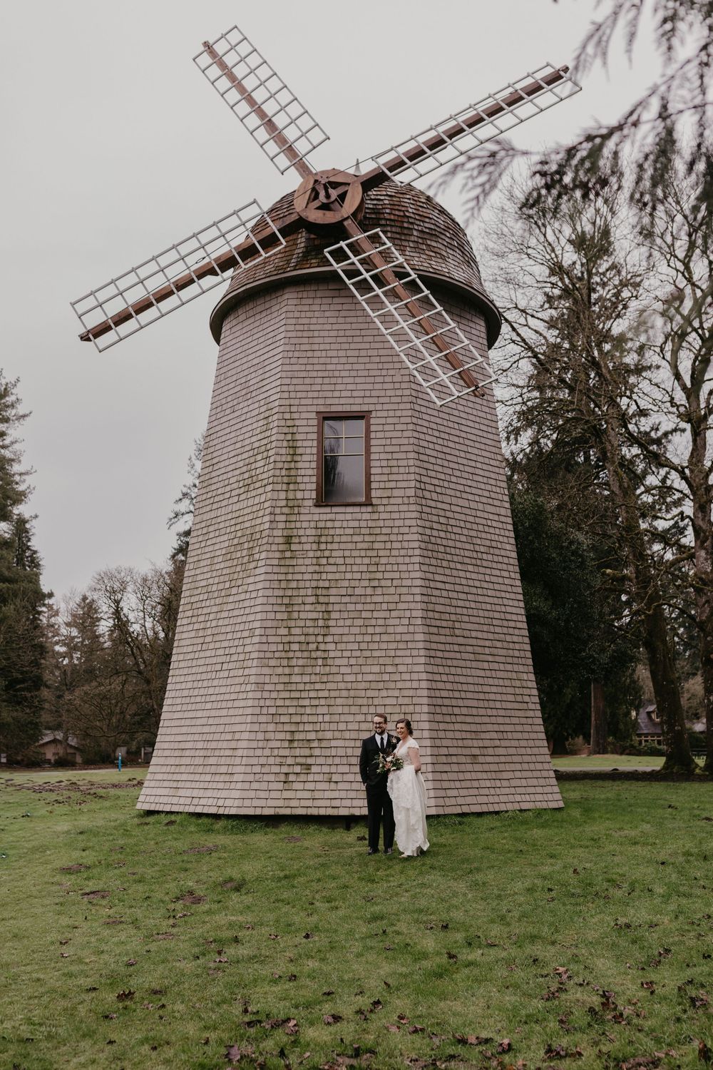 Bride and groom posing together in front of a tall wooden windmill on their wedding day.