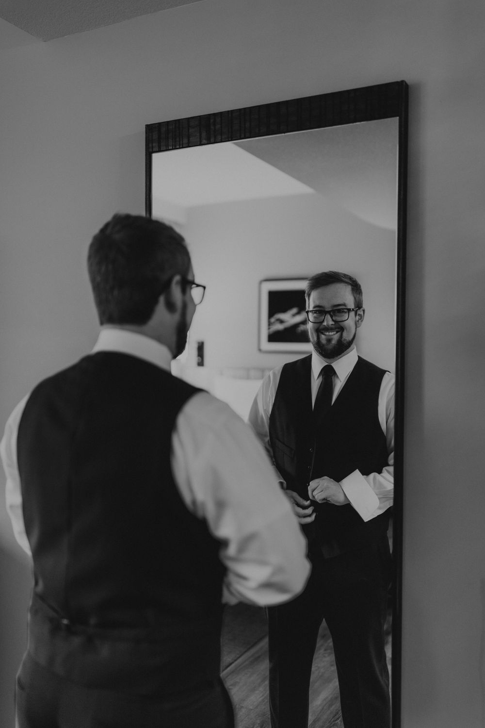 Groom in black vest and tie smiling at his reflection in a full-length mirror before wedding.