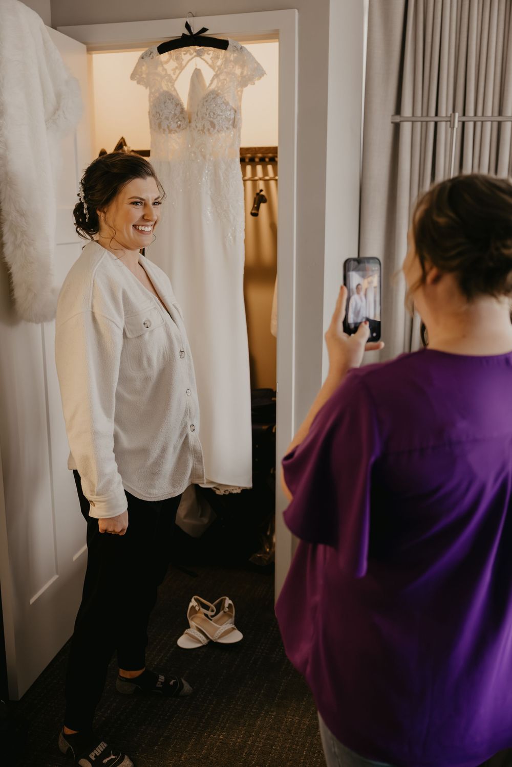 Bride smiling in white shirt as bridesmaid photographs her before the wedding ceremony.
