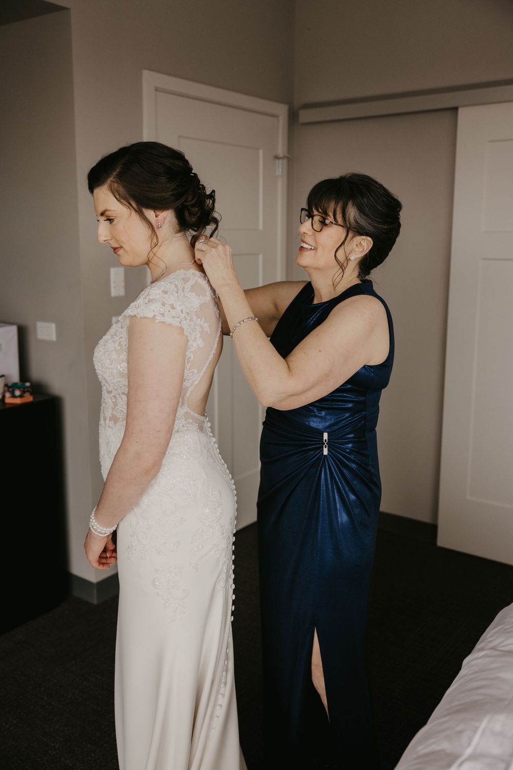 Mother helping bride fasten her lace wedding dress in a hotel room before the ceremony.